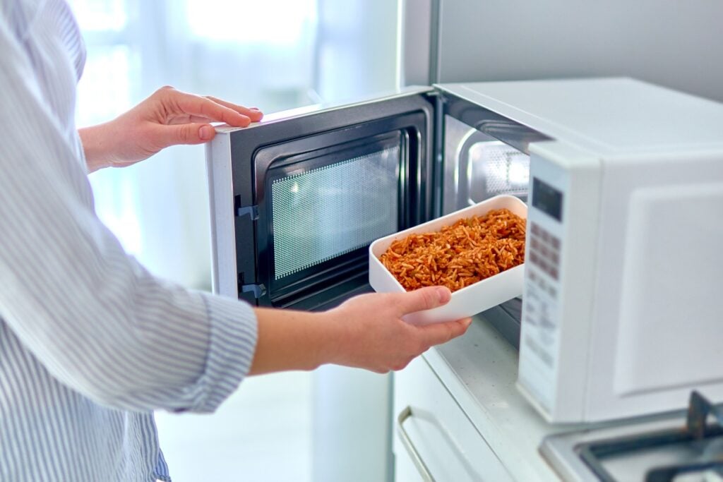 A person places a white container with pasta into a microwave oven. The kitchen appears bright and modern, and the person is wearing a striped shirt.