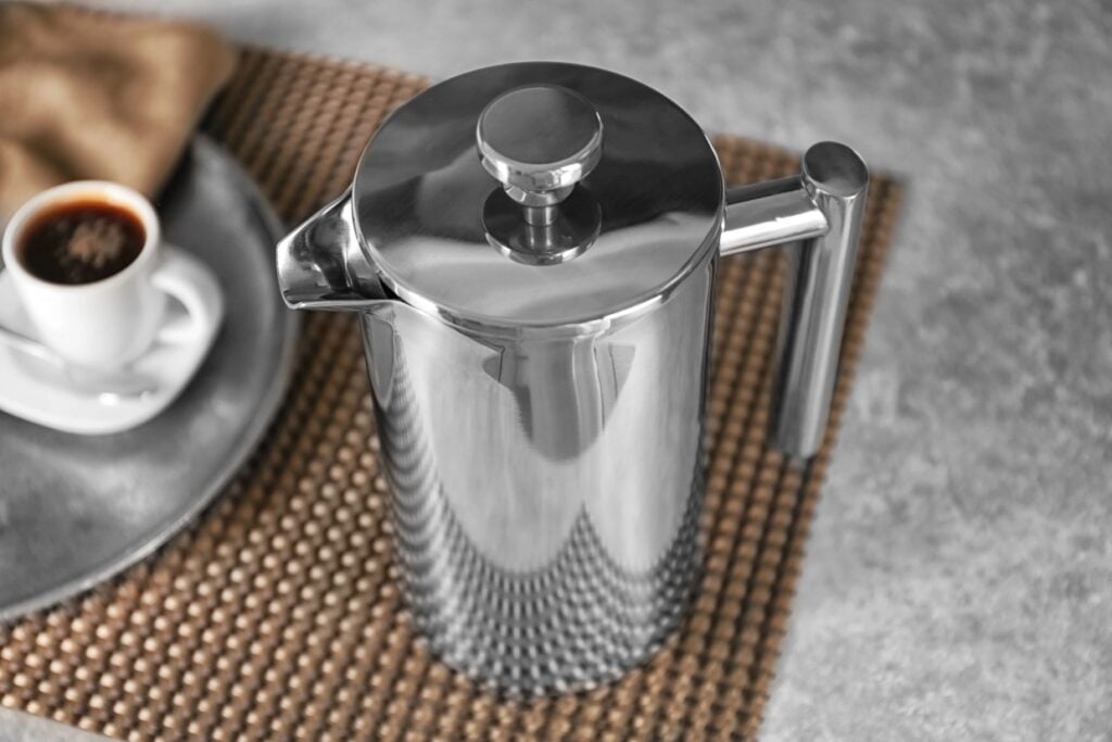 A stainless steel French press sits on a textured placemat next to a small cup of espresso on a saucer. The surface is a gray stone countertop, creating a modern and sleek kitchen setting.