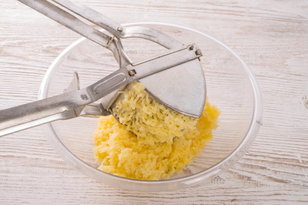 A metal potato ricer is pressing boiled potatoes into a clear glass bowl on a light wooden surface. The riced potatoes form a soft, fluffy pile in the bowl.