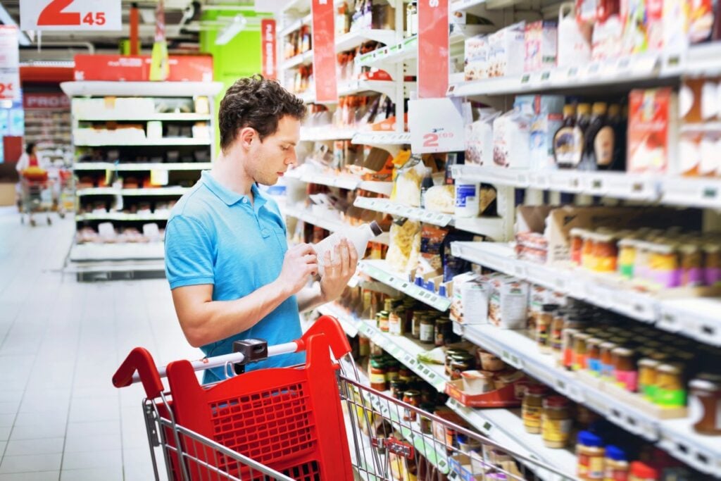 A person in a blue shirt stands in a grocery aisle, examining a product label. A red shopping cart is beside them, filled with items. The shelves are stocked with various food products, sauces, and packaged items. The store is bright and well-lit.