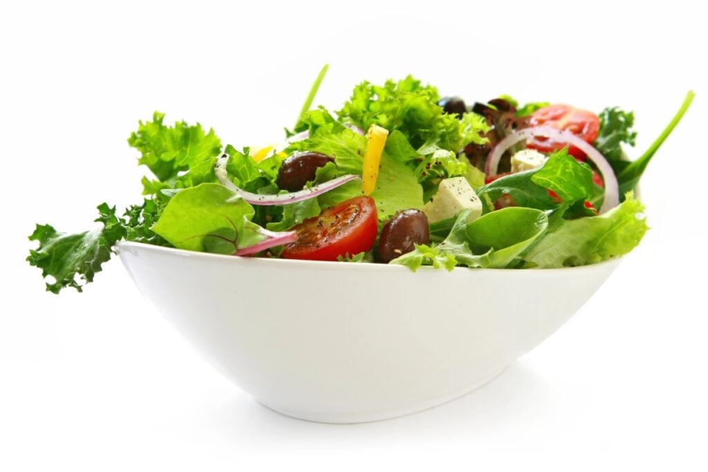 A white bowl filled with a fresh Greek salad, including green lettuce, sliced red onions, cherry tomatoes, black olives, bell peppers, and cubes of feta cheese, all lightly dressed, against a white background.