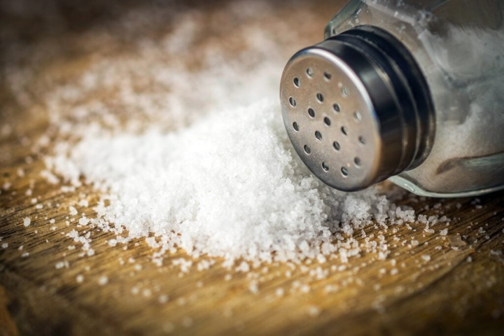 A salt shaker tipped over on a wooden surface, spilling coarse salt in a small pile. The salt grains form a textured pattern against the wood.