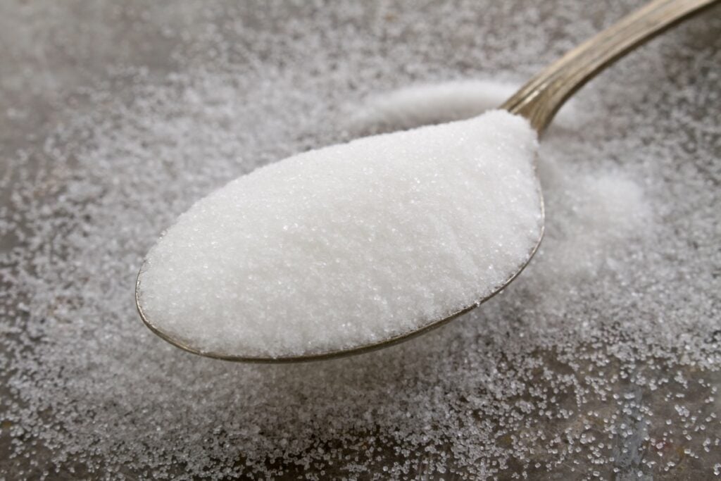 A close-up image of a metal spoon filled with granulated white sugar, resting on a surface sprinkled with additional sugar crystals. The texture of the sugar is clearly visible, showcasing its fine grains.