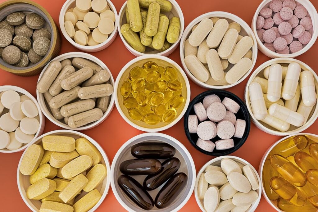 An assortment of various pills and capsules in different shapes and colors displayed in round containers on an orange surface.