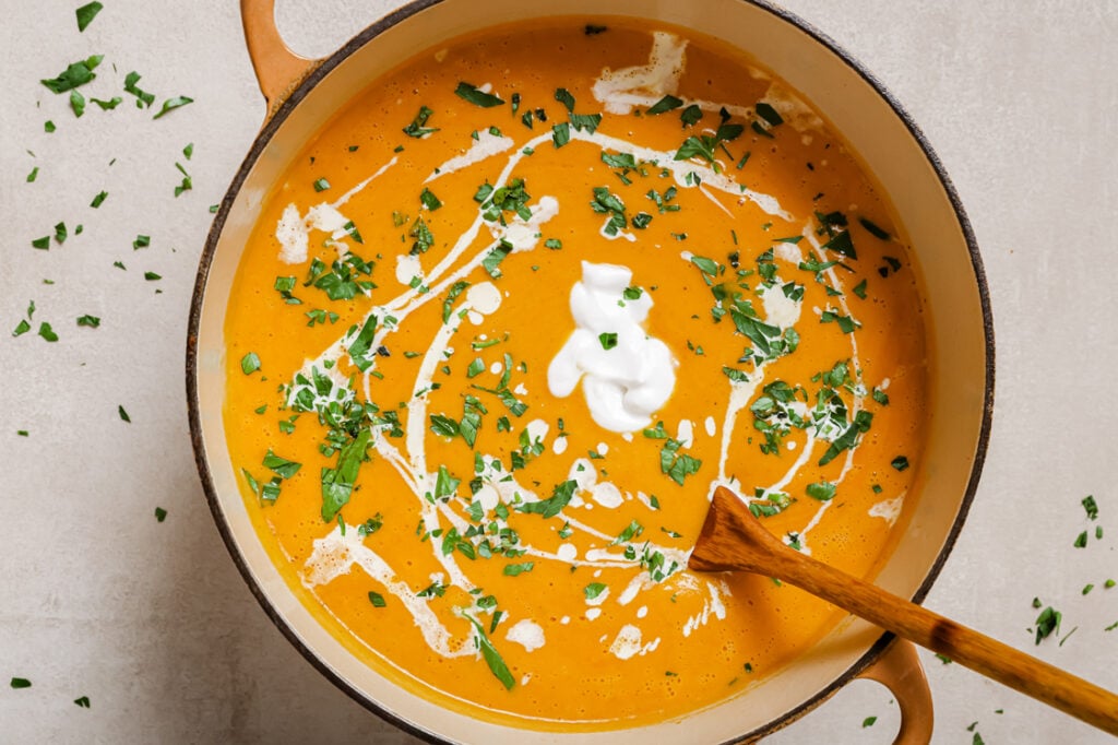 A pot of garnished sweet potato and red pepper soup with a wooden spoon.