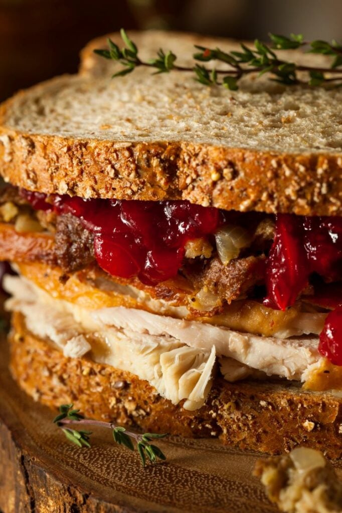 A close-up of a turkey sandwich with layers of sliced turkey, stuffing, and cranberry sauce, all between two slices of grainy whole wheat bread. Sprigs of thyme are garnished on the side.