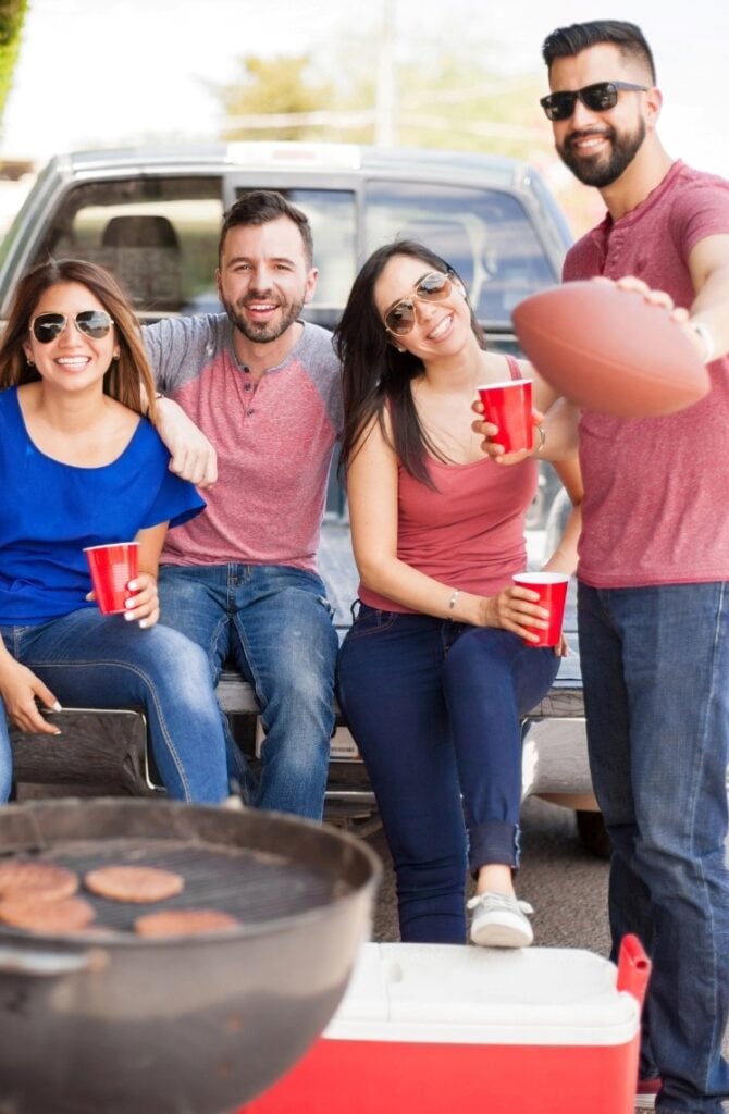 Four friends enjoying a tailgate party by a truck. They are holding red cups and one is holding a football. A grill with burgers is in the foreground, and there's a cooler nearby. They are smiling and wearing casual outfits and sunglasses.