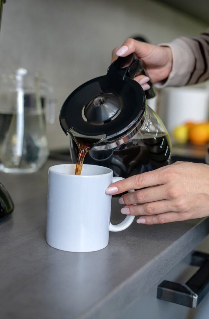 A person pours coffee from a glass carafe into a white mug on a kitchen counter. In the background, there is another glass container filled with water and some fruit slightly out of focus.