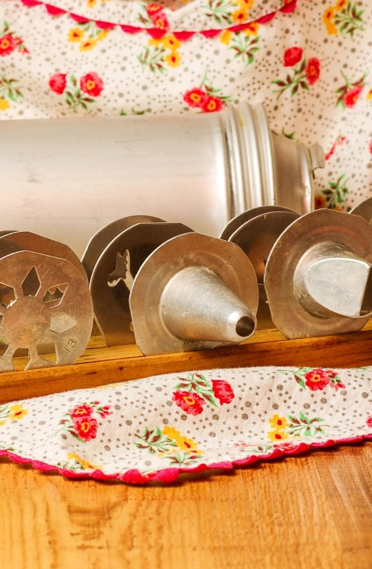 A collection of metal cake decorating tips and presses arranged on a wooden surface, with a floral fabric in the background. The scene evokes a vintage baking atmosphere.