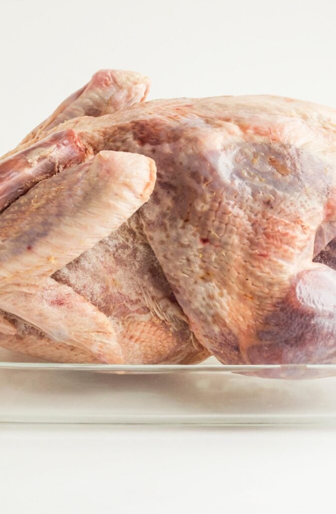 A raw whole turkey placed in a clear glass dish, ready for cooking. The turkey's textured skin is visible under bright lighting against a neutral background.