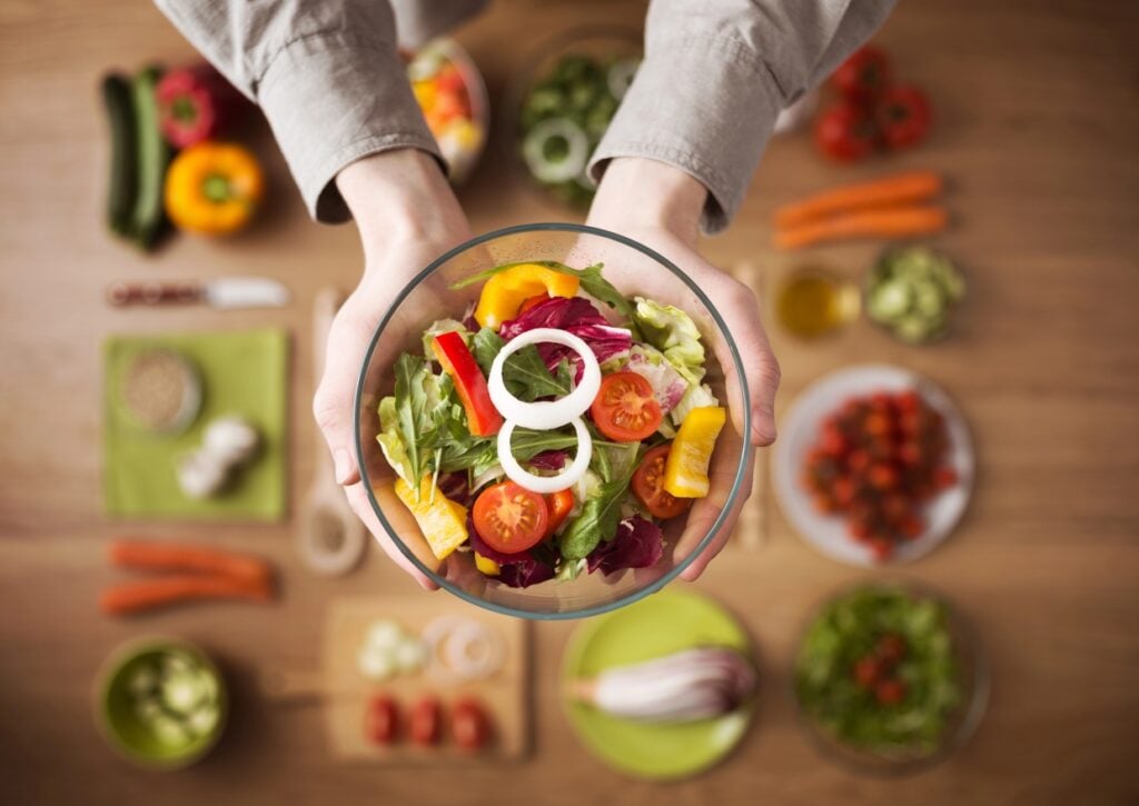 A person holding a bowl of fresh salad with lettuce, cherry tomatoes, and bell peppers. In the background, there's a wooden table with various vegetables like carrots, cucumbers, and peppers, along with bowls of ingredients.