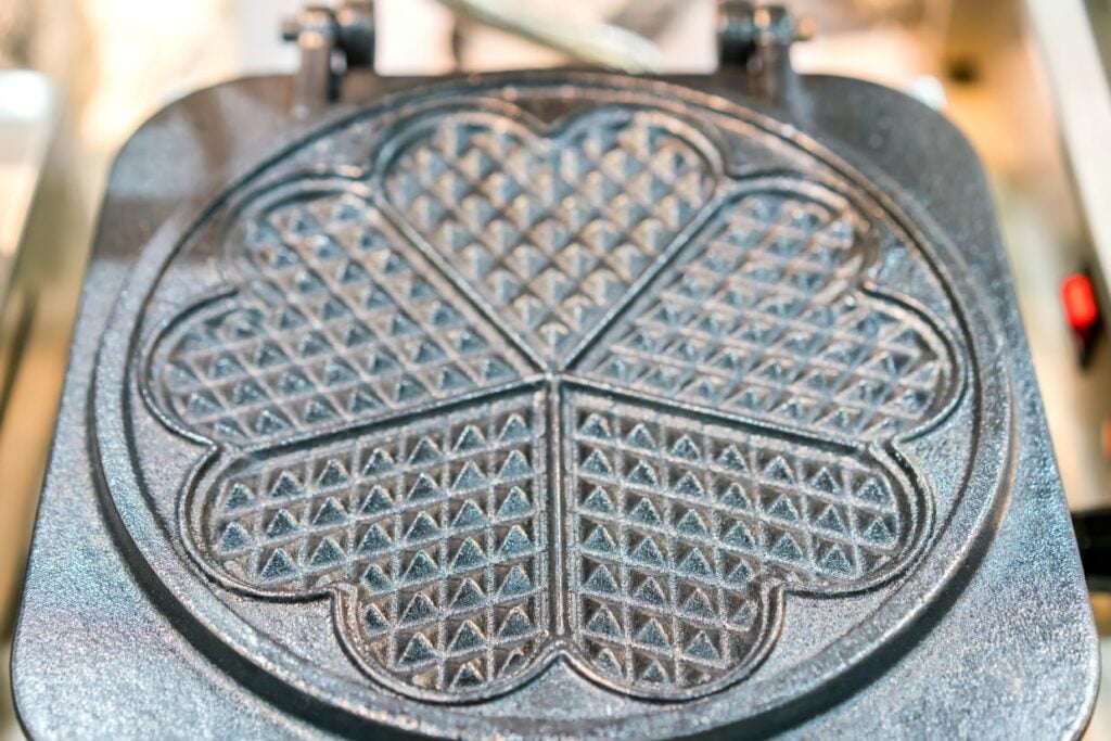 Close-up of a waffle iron plate with a heart-shaped pattern divided into five segments, each featuring a triangular grid design. The metal surface is shiny and textured, set in a kitchen environment.