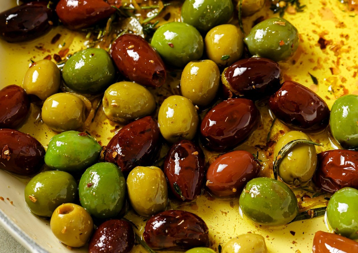 A close-up of assorted green and brown olives marinated in oil and herbs. The olives are glossy and surrounded by visible herbs and spice flakes.