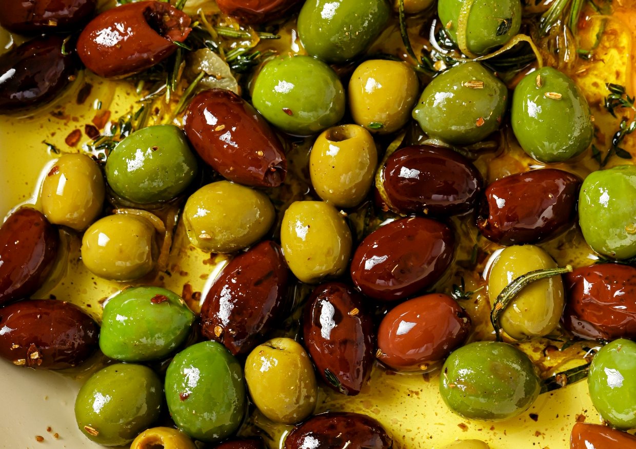 A close-up of assorted green and brown olives marinated with herbs and spices in olive oil. The olives vary in size and color, glistening under the light, surrounded by sprigs of rosemary and scattered chili flakes.