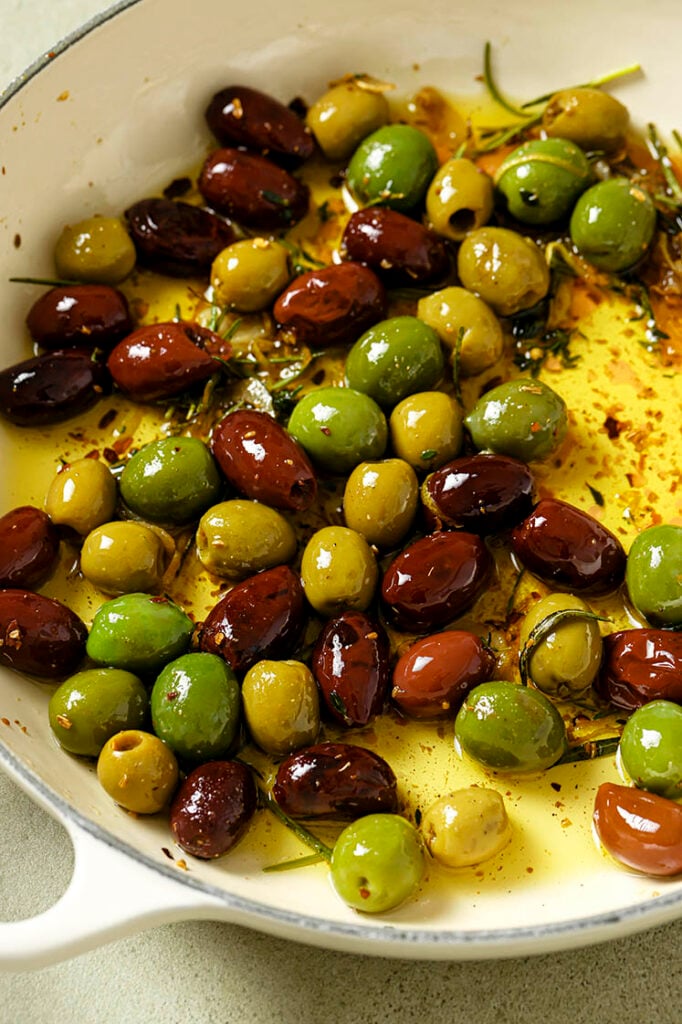 A skillet filled with warm marinated olives, garnished with herbs and garlic.