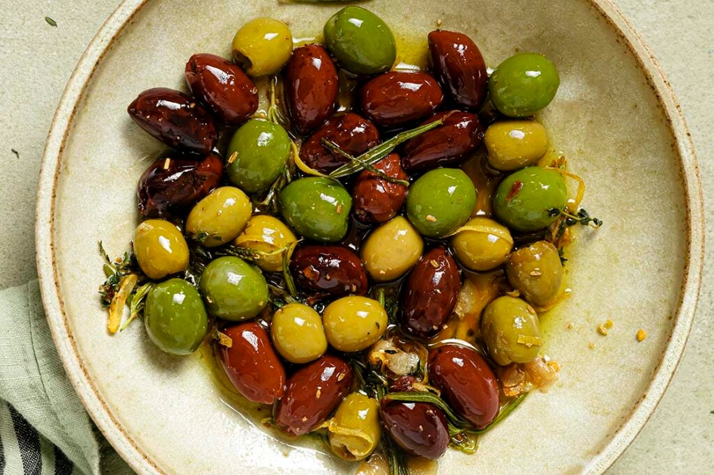 A ceramic bowl filled with warm marinated olives, garnished with herbs and garlic.