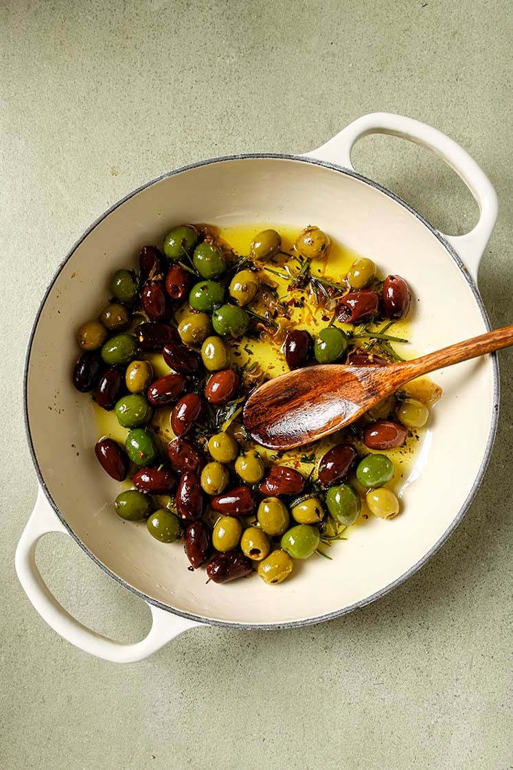 A skillet pot filled with a mix of green and black olives, simmered in olive oil, garlic, spices, and lemon zest using a wooden spoon.