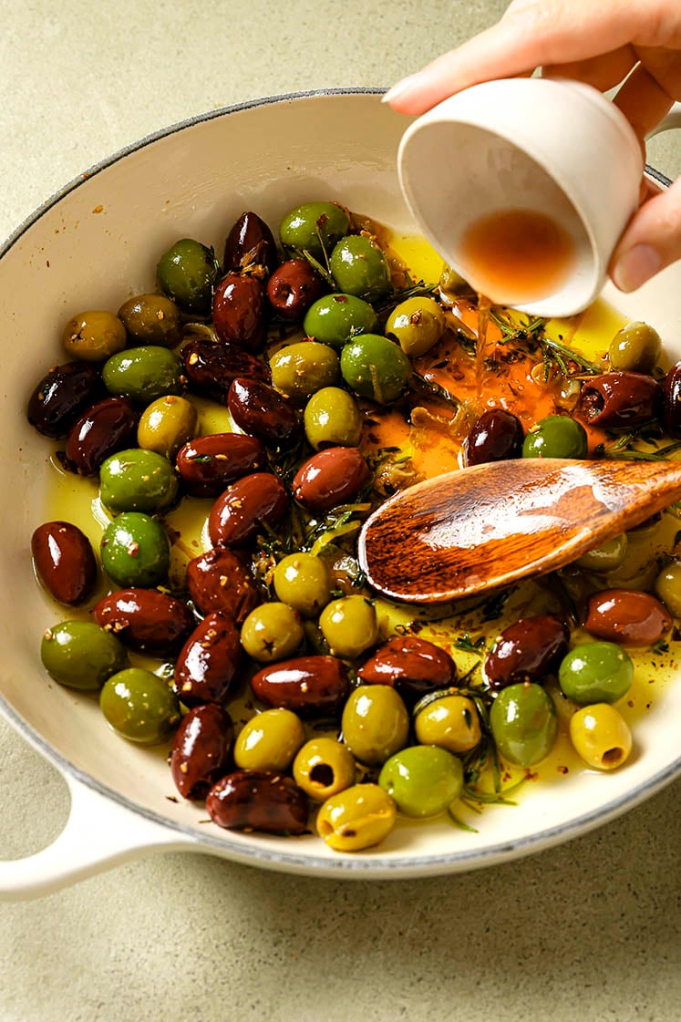 Red wine vinegar is being poured on a skillet pot filled with a mix of green and black olives, simmered in olive oil, garlic, spices, and lemon zest with a wooden spoon.