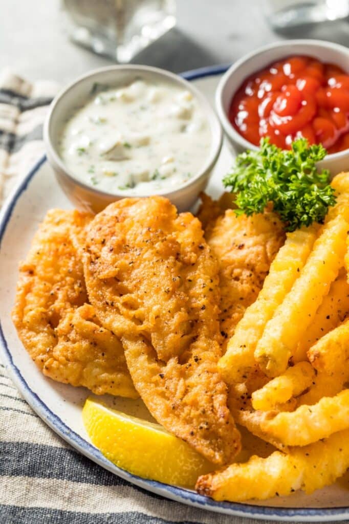 A plate of golden-brown fried fish and crinkle-cut fries, accompanied by small bowls of tartar sauce and ketchup. A lemon wedge and a sprig of parsley garnish the dish, which rests on a striped napkin.