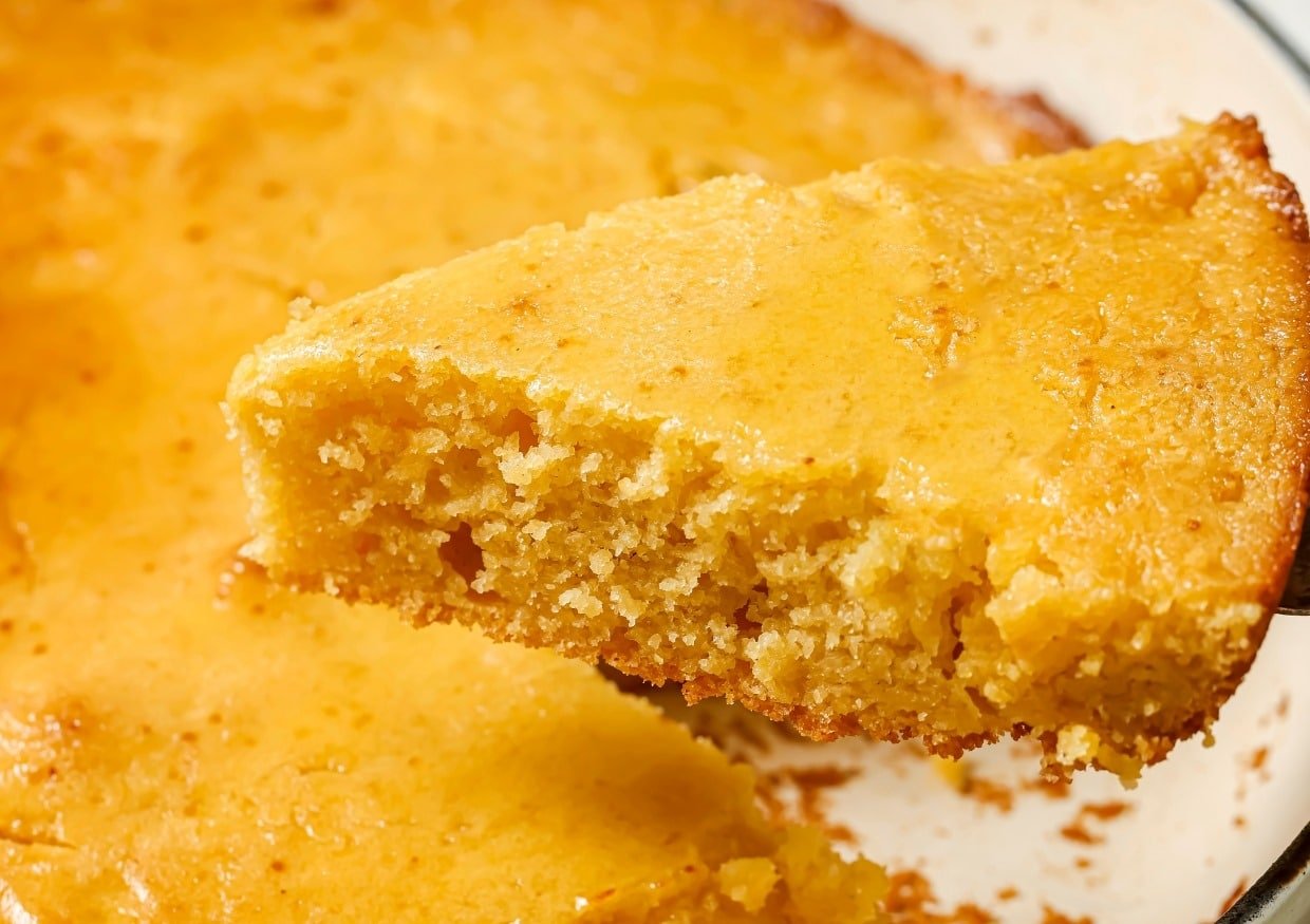 A close-up of a slice of cornbread being lifted from a round pan. The cornbread has a golden-brown crust and a moist, crumbly interior.