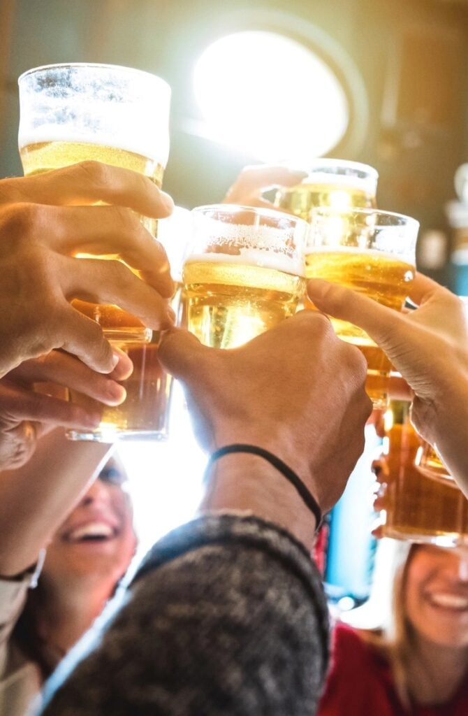 A group of people raising beer glasses in a toast, with smiling faces visible beneath the glasses. The setting appears to be a lively indoor gathering with natural light streaming in from a window behind them.