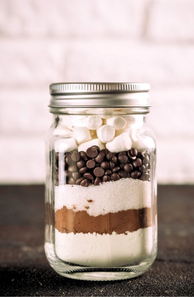 A glass jar filled with layers of ingredients for hot chocolate, including marshmallows, chocolate chips, cocoa powder, sugar, and flour. The jar is sealed with a metal lid and set against a blurred white brick background.