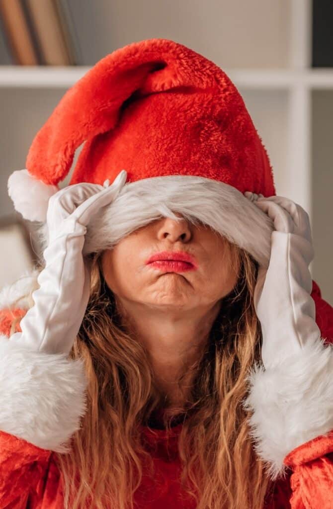 A person in a red Santa outfit, including a hat and white gloves, playfully pulling the hat down over their eyes. They have long hair and are posing with a lighthearted expression.