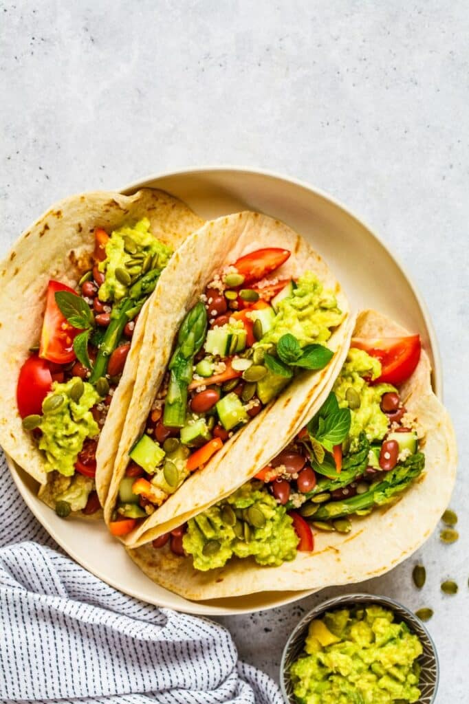 A plate with two colorful vegan tacos filled with avocado, tomatoes, quinoa, asparagus, beans, and seeds, garnished with fresh basil leaves. A small bowl of guacamole is placed beside the plate. A textured cloth is partially visible.