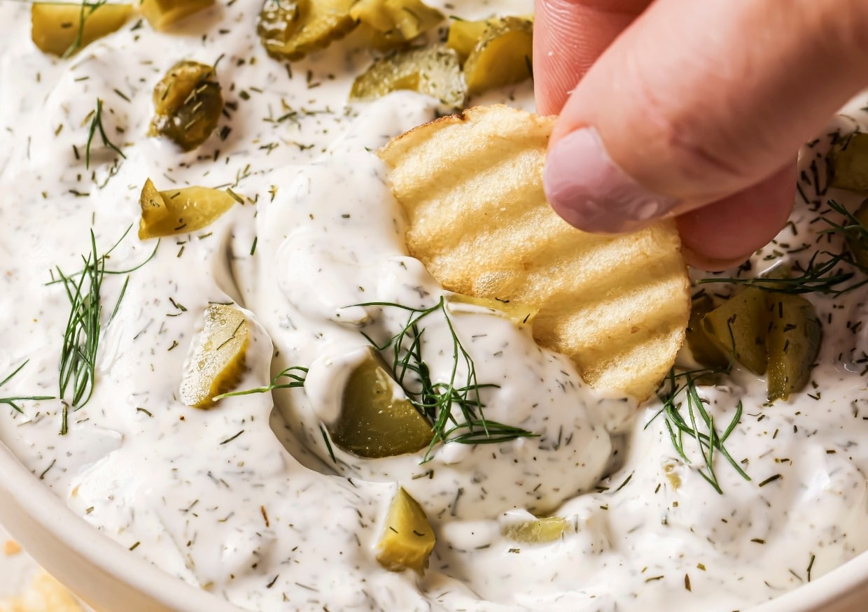 A hand dipping a ridged potato chip into a creamy dill dip garnished with chopped pickles and fresh dill sprigs.