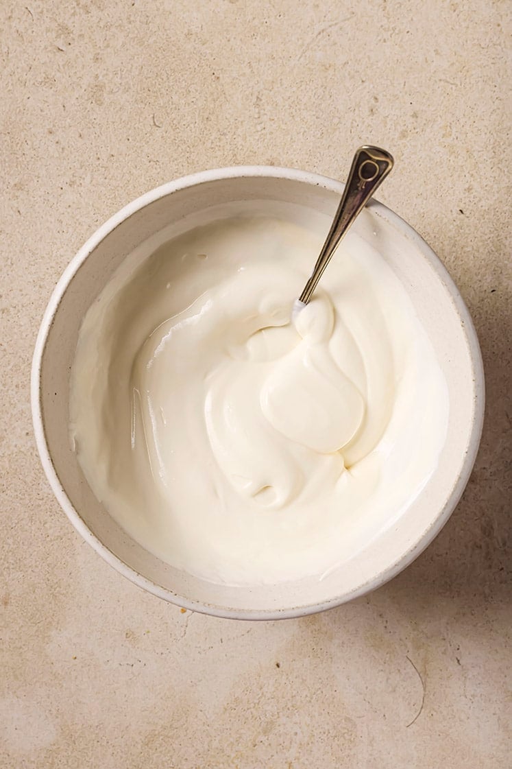 A bowl of creamy white yogurt with a spoon in it, placed on a light-colored textured surface.