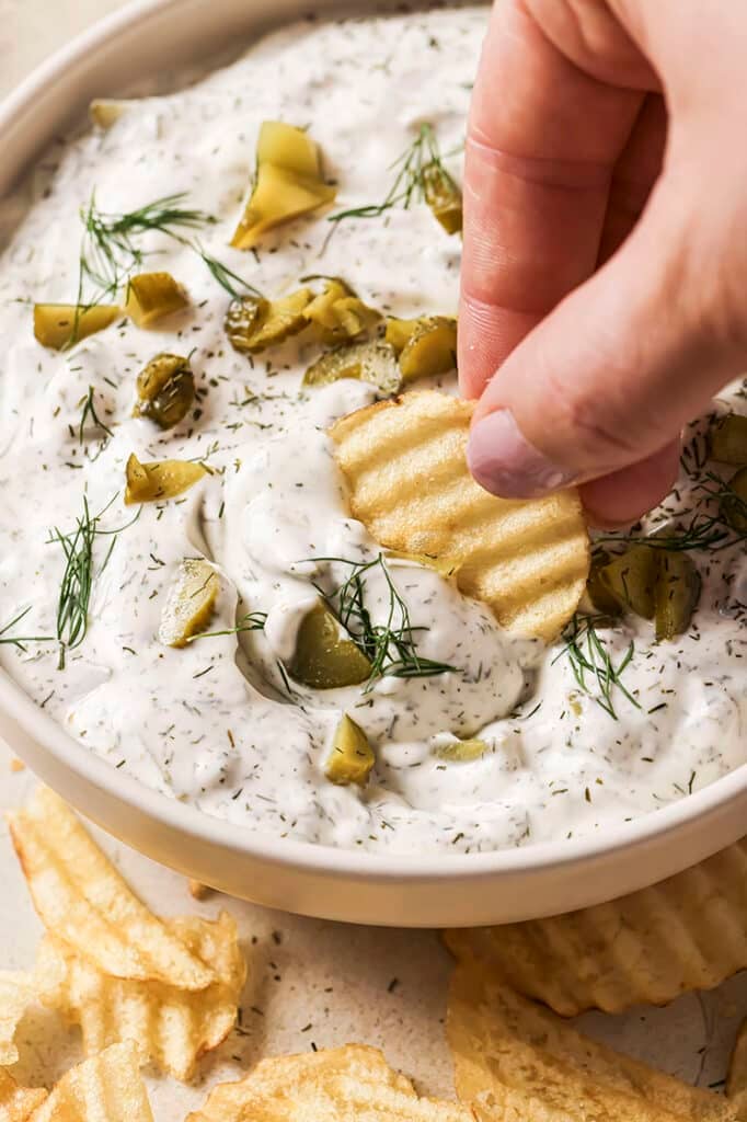 A hand dips a crinkle-cut potato chip into a bowl of creamy dill pickle dip. The dip is garnished with chopped pickles and fresh dill, surrounded by additional chips on the side.