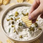 A hand dipping a ridged potato chip into a bowl of creamy dill pickle dip, garnished with chopped pickles and fresh dill. The bowl is surrounded by scattered potato chips on a light-colored surface.