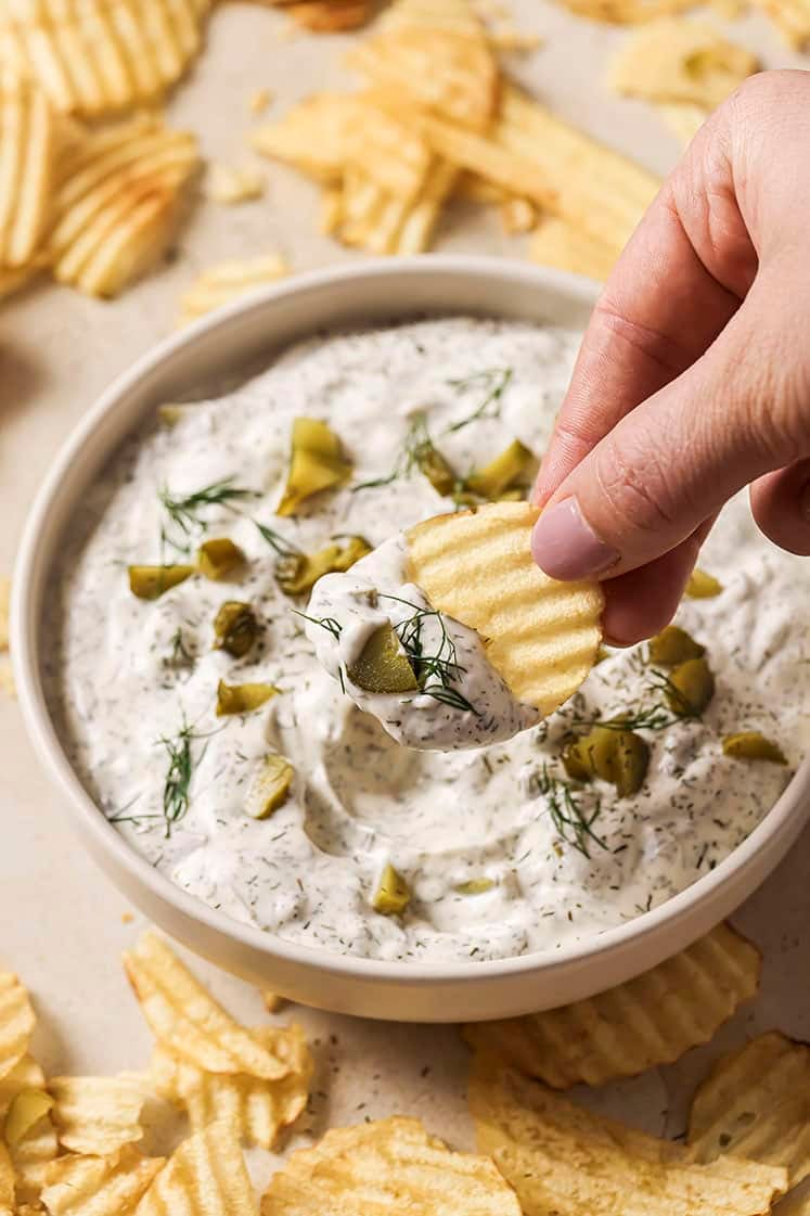 A hand dipping a ridged potato chip into a bowl of creamy dill pickle dip, garnished with chopped pickles and fresh dill. The bowl is surrounded by scattered potato chips on a light-colored surface.