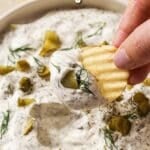 Close-up of a hand dipping a wavy chip into a bowl of creamy dill pickle dip garnished with chopped pickles and dill. The text reads "Easy Dill Pickle Dip" at the top of the image. Crinkle-cut chips are scattered around the bowl.