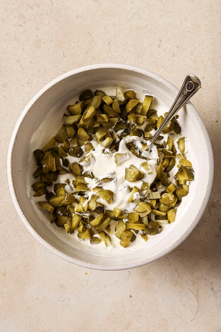 A bowl of chopped pickles mixed with yogurt, featuring a silver spoon, on a light beige surface.