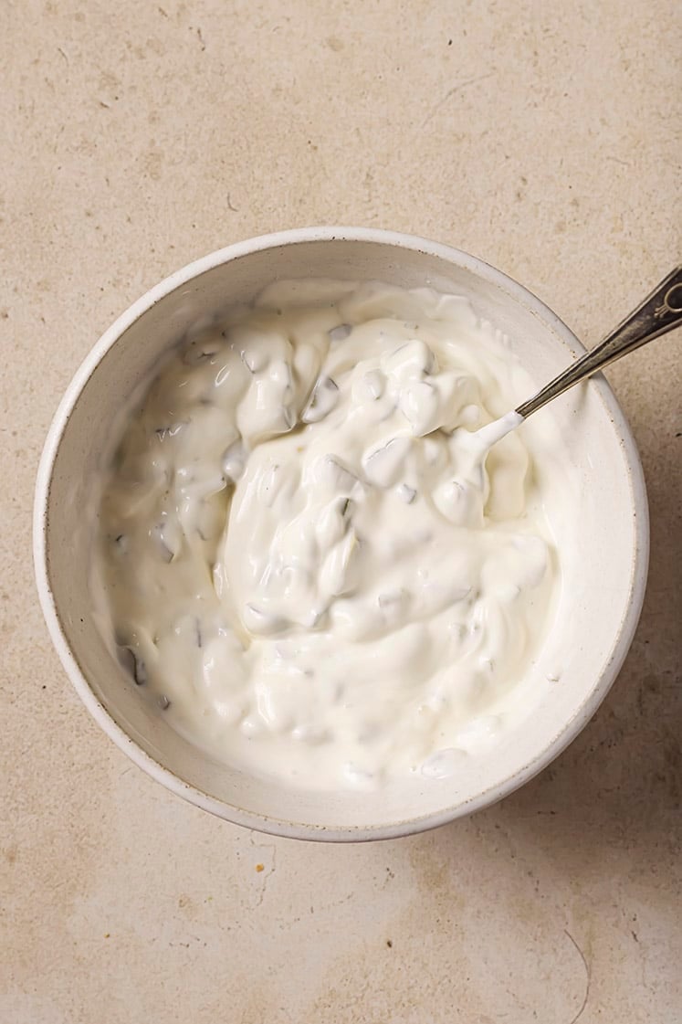 A bowl of creamy white dip with visible chunks with a spoon resting inside. The bowl sits on a beige textured surface.