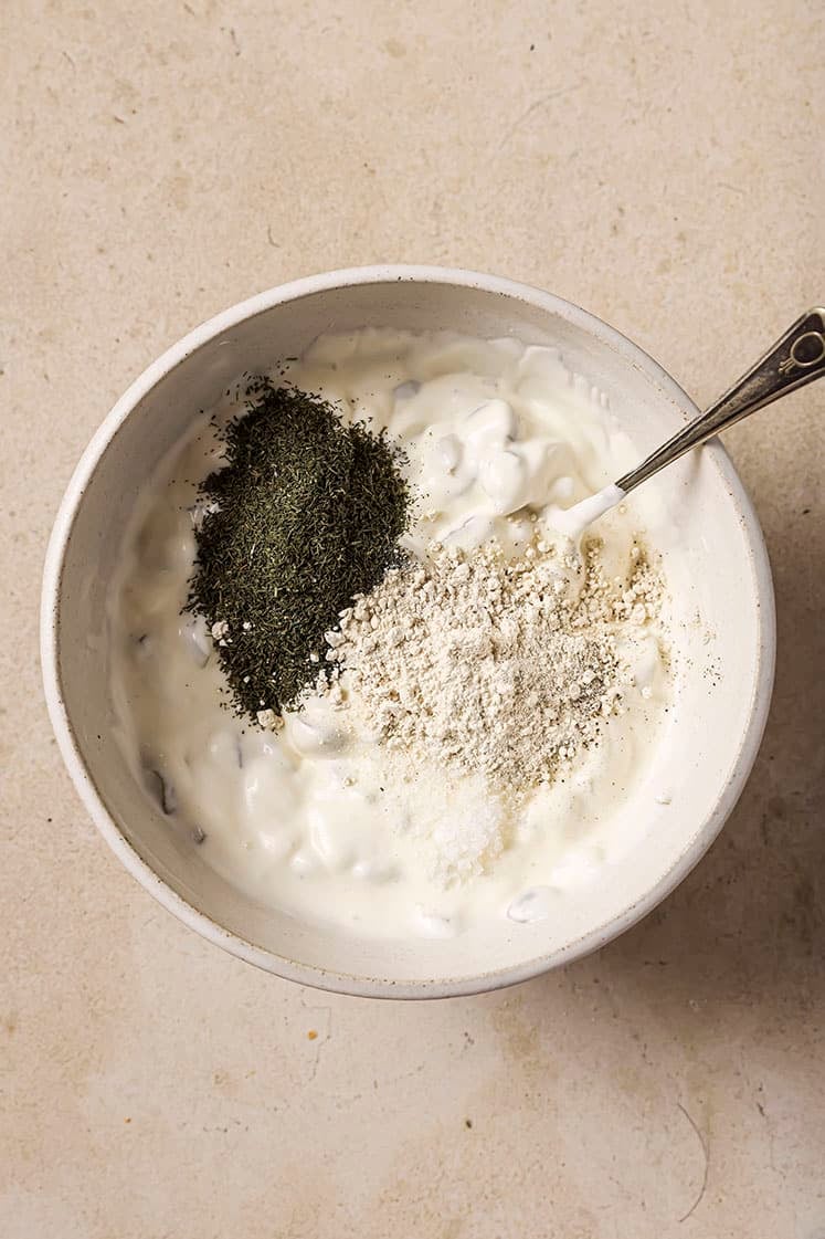 A bowl of creamy sauce contains a spoon and is topped with green herbs and a pile of white seasoning. The mixture rests on a light-colored, textured surface.