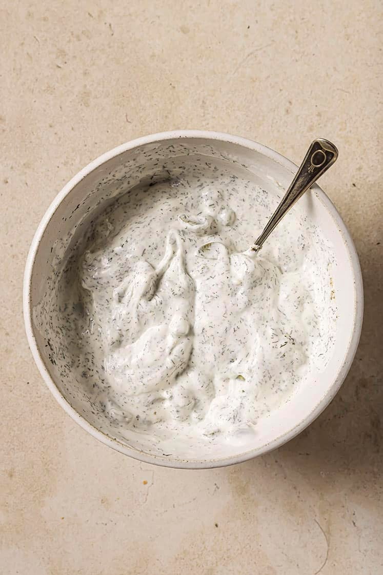 A bowl of creamy, white dip speckled with fine green herbs, with a spoon resting inside, placed on a light beige surface.