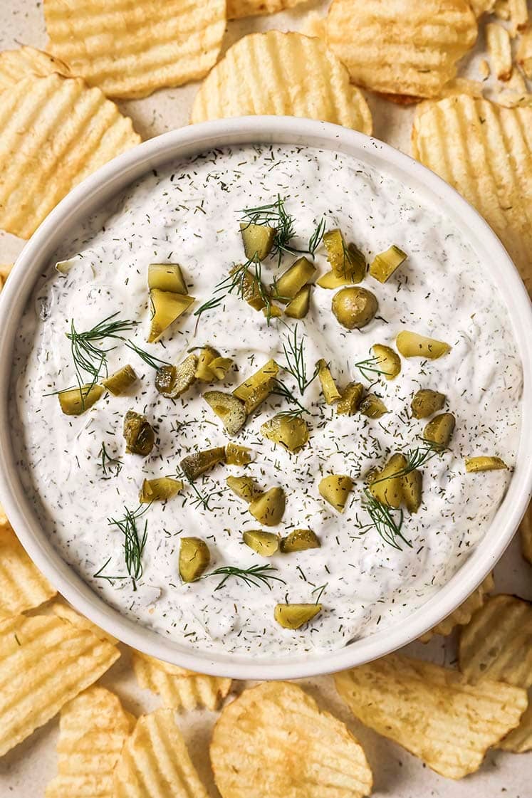 A bowl of creamy dip topped with chopped pickles and dill, surrounded by ridged potato chips.