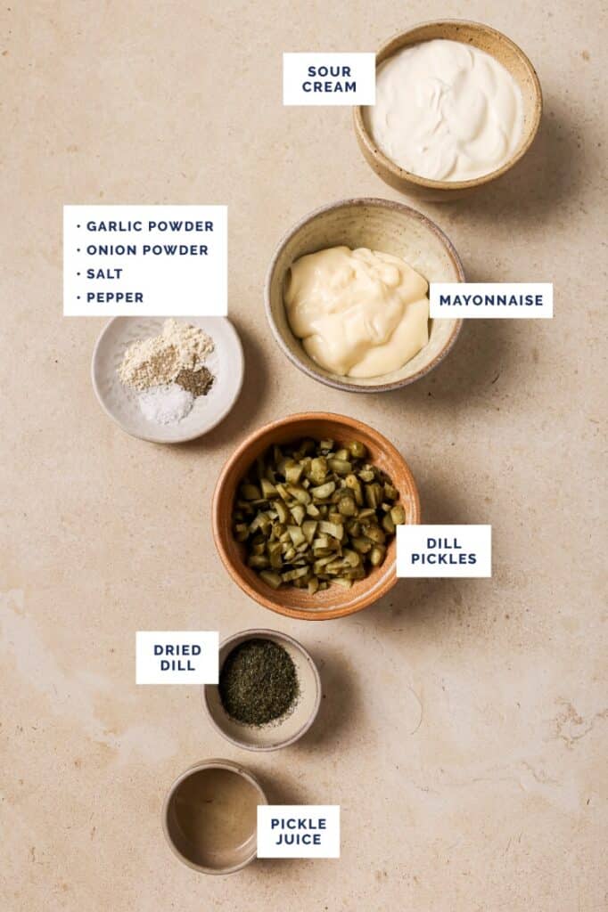 Ingredients for a recipe arranged on a beige surface: bowls of sour cream, mayonnaise, diced dill pickles, dried dill, and pickle juice. A small plate contains garlic powder, onion powder, salt, and pepper.