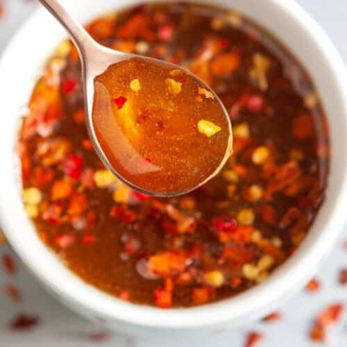 A close-up of a spoon holding a spicy, reddish-brown sauce with visible chili flakes, above a white bowl filled with the same sauce. Some chili flakes are scattered around, enhancing the spicy appearance.