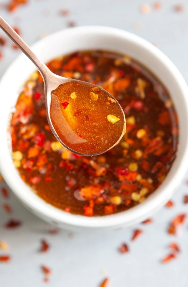 A close-up of a spoon holding a spicy, reddish-brown sauce with visible chili flakes, above a white bowl filled with the same sauce. Some chili flakes are scattered around, enhancing the spicy appearance.