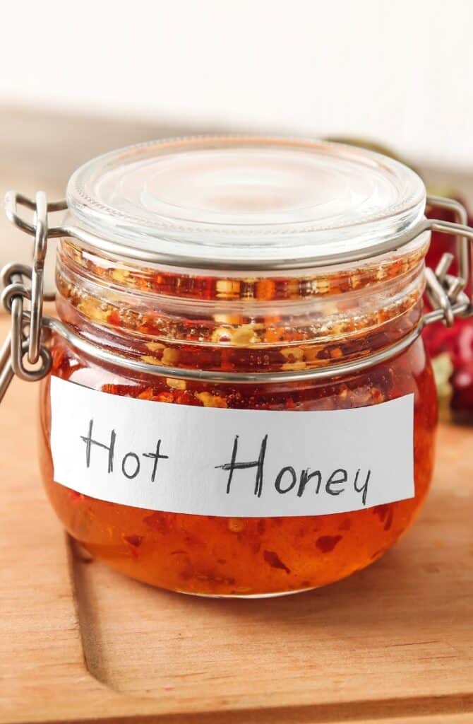 A glass jar of hot honey with a metal clasp sits on a wooden surface. The jar has a handwritten label that reads "Hot Honey," and the honey inside appears infused with chili flakes.