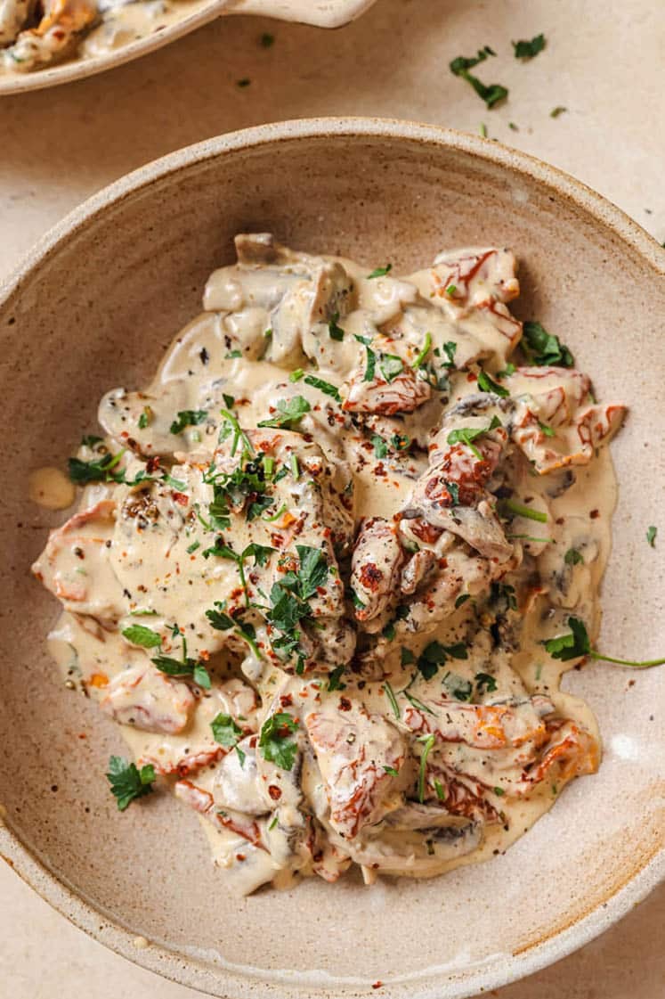 A bowl filled with creamy sun-dried tomato and mushroom sauce, garnished with fresh parsley. The dish is served in a rustic stoneware bowl, and the sauce has a rich, creamy texture with visible slices of tomatoes and mushrooms.