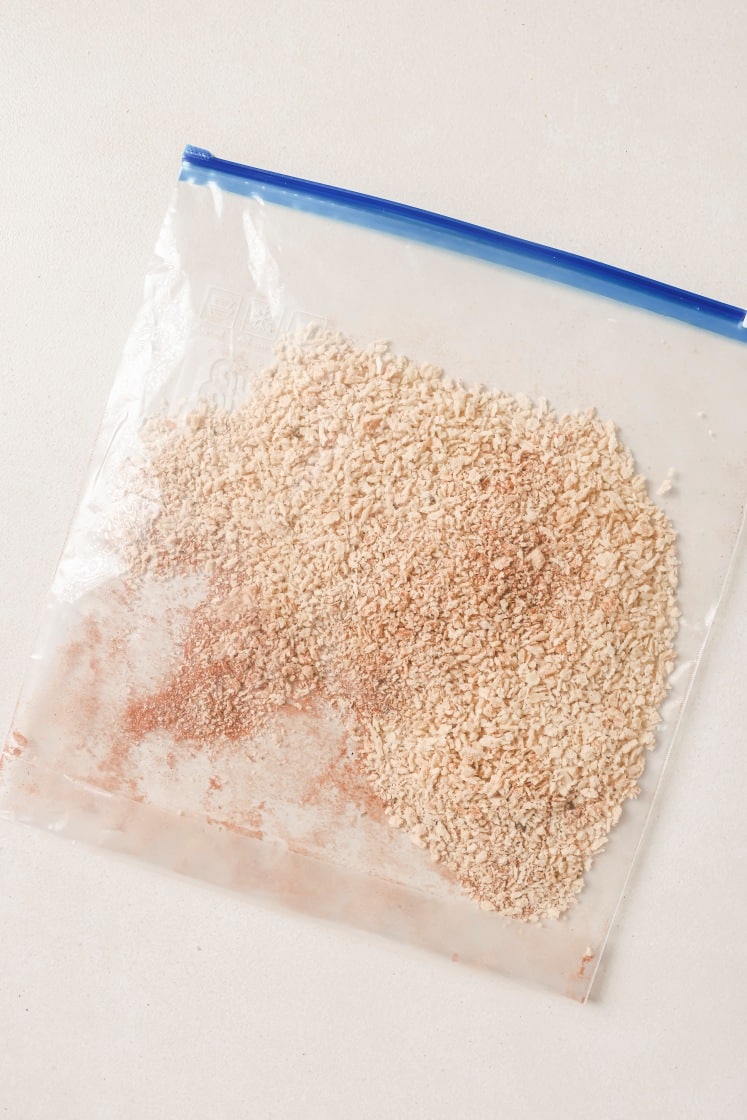 A transparent zip-top bag containing a mix of fine, beige particles and a hint of reddish powder. The bag is lying flat on a light surface.