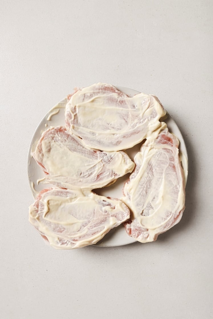 Four raw pork chops coated with white mayonnaise are arranged on a round white plate against a light background.