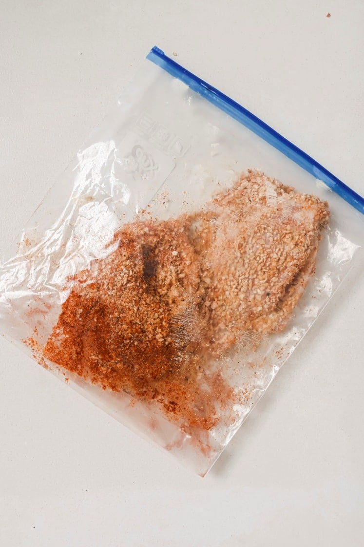 A clear resealable plastic bag containing a breaded pork chops and a mixture of breadcrumbs and spices. The bag is placed on a white surface.