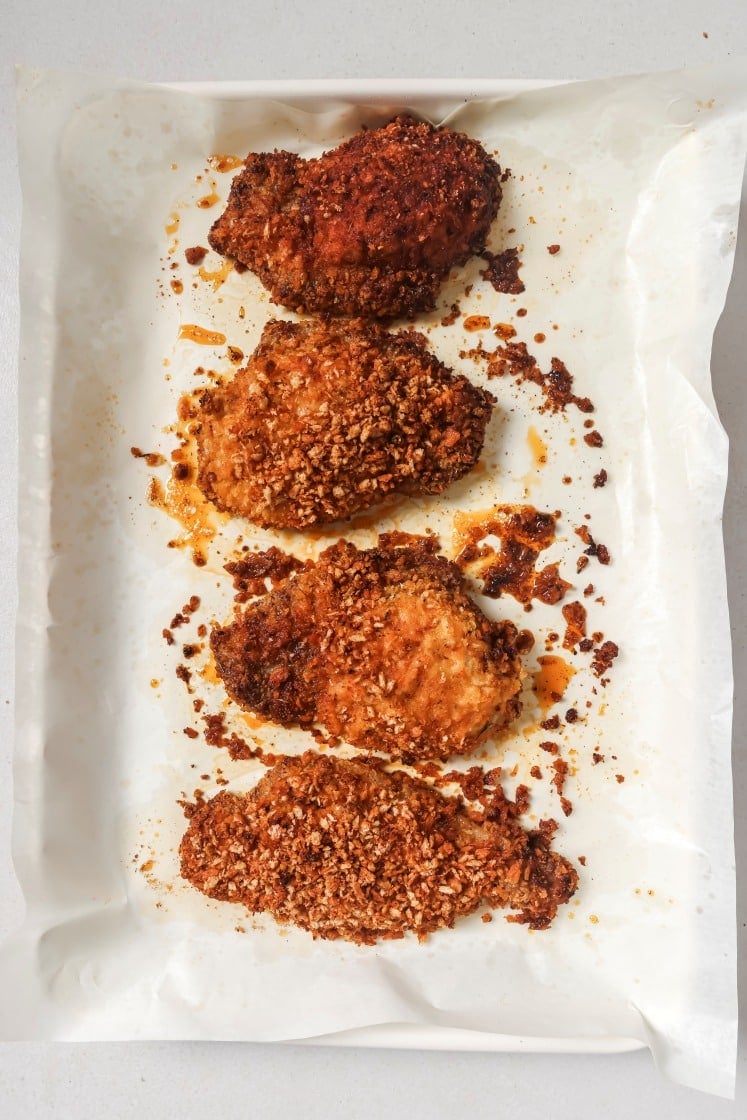 Four pieces of crispy, golden-brown pork chops on a parchment-lined tray. The chicken has a textured, spiced coating with bits scattered around, and a slight oily sheen is visible.