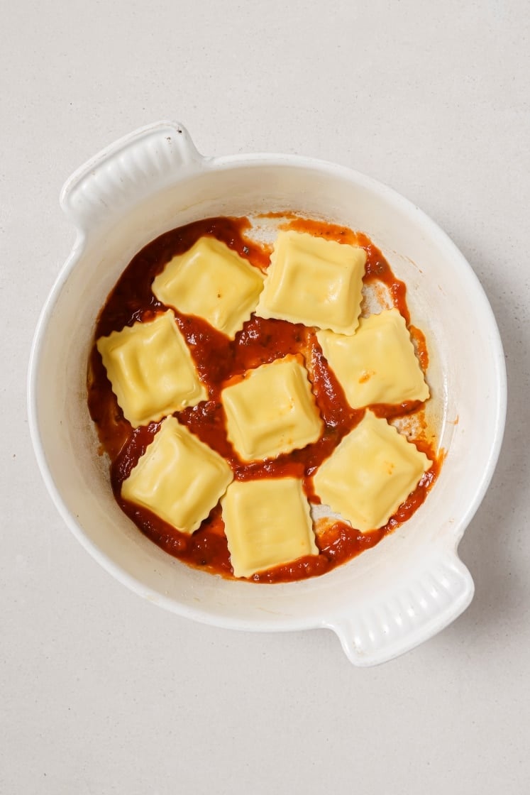 White dish with eight pieces of uncooked ravioli placed over a layer of tomato sauce. The dish is on a light-colored surface.