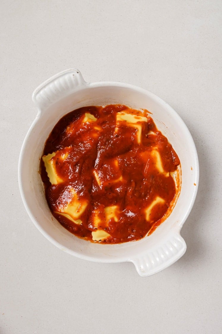 White ceramic dish filled with ravioli covered in tomato sauce on a light surface.